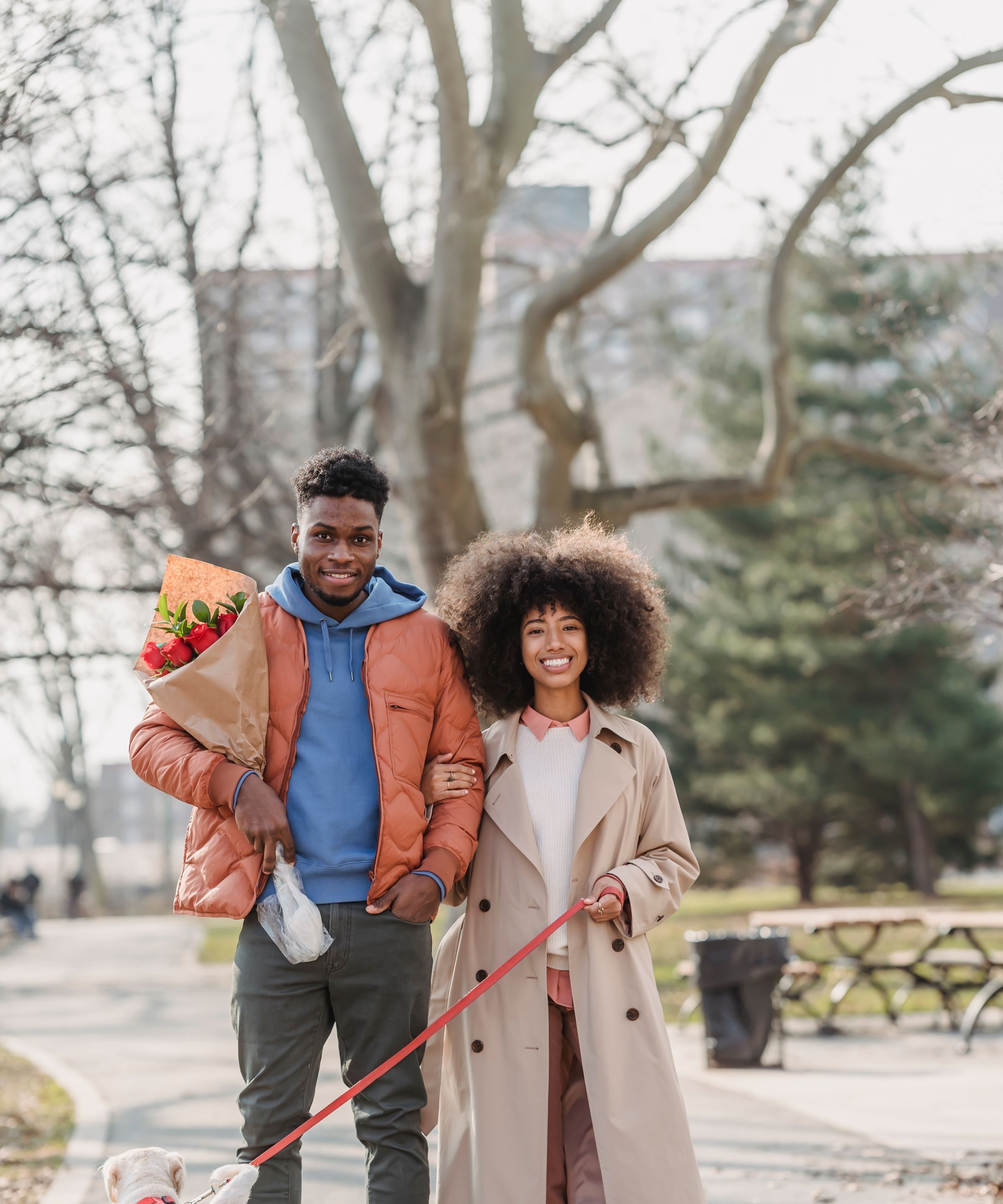 A young couple arm in arm in a park, flowers in hand, walking their dog